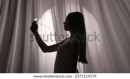 Medium side view shot of a young woman silhouette wrapped in a towel using a facial mist sprayer, moisturizing device on her skin in a muffled light. Royalty-Free Stock Photo #2371119579