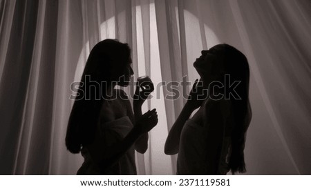 Medium side view shot of two sisters, young women's silhouettes wrapped in a towel. One is holding a jar of body cream, the other one in touching her skin in a muffled light. Royalty-Free Stock Photo #2371119581