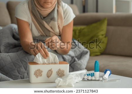 Medicines for colds and paper napkins are on the table, a woman with signs of flu and colds is sitting in the background and takes a napkin out of the box. Focus on medications Royalty-Free Stock Photo #2371127843