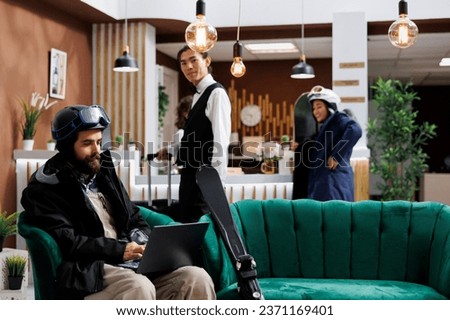 Caucasian man utilizing laptop to book activities for winter vacation at ski resort. Male guest dressed in snow clothing using minicomputer in lounge area while employee takes suitcase to hotel room.