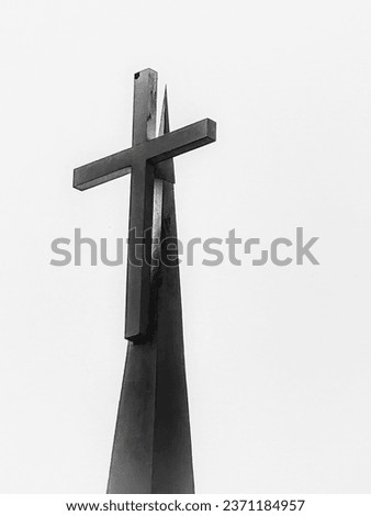A black and white picture of a cross with a pointed vertical support behind it. A strong light is showing on the support behind the cross. 