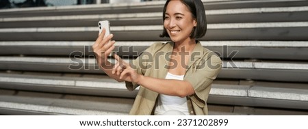Portrait of asian beautiful girl takes selfie on smartphone. Young korean woman sits on stairs outdoors and makes photos on mobile phone.