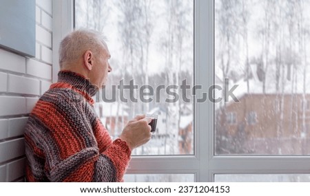 A man dressed in a knitted warm sweater stands at the window with a cup of mulled wine in his hand