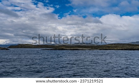 Rocky islets with sparse stunted vegetation are visible in the Beagle Channel. A mountain range against a background of blue sky and clouds in the distance. Ripples on the water. Argentina.  