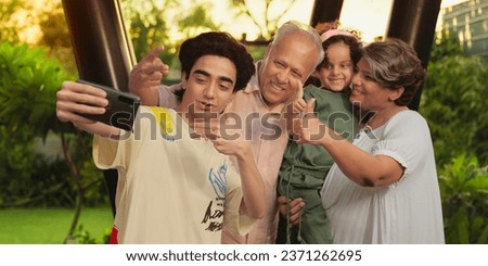 Happy Indian smiling older family standing outdoor home taking selfie enjoy summer vacation together. Grandparents recording video with little girl child making fun spend weekend holiday time at house
