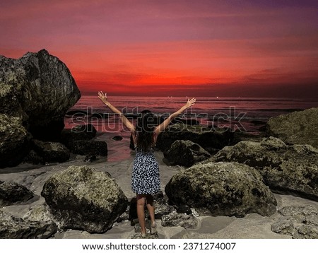 A beautiful woman in a flower dress stands by the ocean's edge in stones Bali, her hands outstretched, embracing the enchanting sunset. The vibrant colors of the sky.
