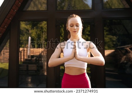 Portrait of peaceful redhead female putting hands in namaste mudra posture standing with closed eyes on background of country house. Front view of calm young woman practicing yoga alone outdoors.