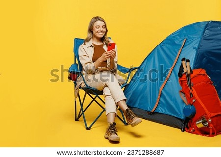 Full body smiling young woman sit near bag stuff tent use mobile cell phone isolated on plain yellow background. Tourist leads active lifestyle walk on spare time. Hiking trek rest travel trip concept