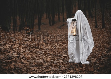Spooky ghost holding glowing lantern in moody dark autumn forest. Person dressed in white sheet as ghost with light in evening fall woods. Happy Halloween! Trick or treat