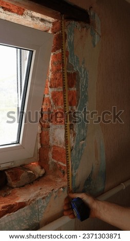 a vertical image where a builder, invisible in the frame, uses a tape measure to measure the height of a window opening in the process of installing a new small window in an old room