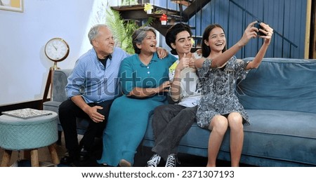 Happy Indian excited family sitting on sofa smiling teenage girl hold smartphone taking selfie at house. Old grandparents couple posing for funny photo or recording video blog enjoy weekend together