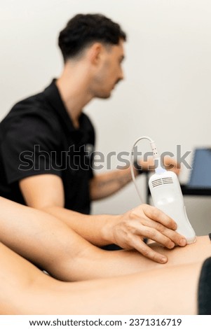A male physical therapist is performing an ultrasound on a patient's femoral to detect pain. The probe in the client's leg is in the foreground. Concept of ultrasound in physiotherapy clinics. Royalty-Free Stock Photo #2371316719