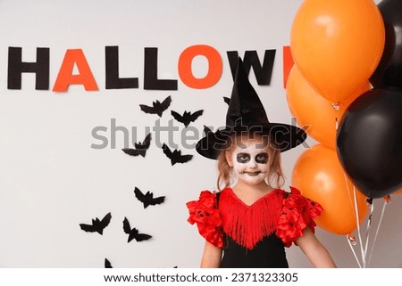 Little girl dressed for Halloween as witch with balloons and decor near light wall