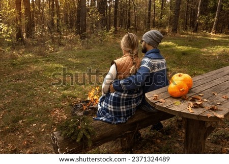 Man and woman sit by fire in autumn forest on benches at table with pumpkins. Family on picnic relax outdoors. Leisure, weekend in nature