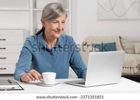 Beautiful senior woman with cup of drink using laptop at white table indoors