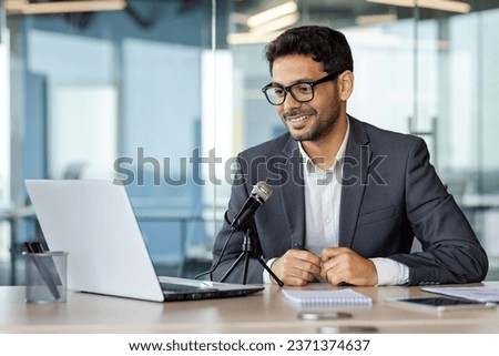 Young successful business coach recording podcast audio course about success and money, man using professional microphone and laptop while sitting at desk in middle of office.