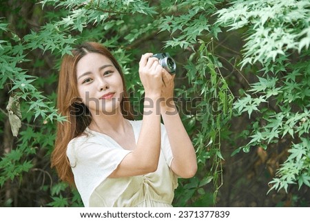 A beautiful Asian woman in a dress is taking pictures with a camera in a forested park during the summer day.	