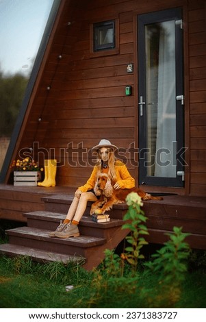 a long-haired blonde European teen girl in a mustard sweater and hat is sitting on the steps of a wooden house next to an English Cocker spaniel dog on an autumn day Royalty-Free Stock Photo #2371383727