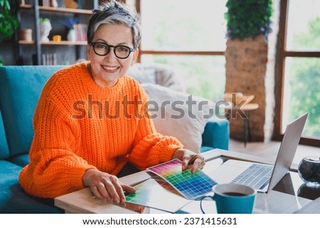 Photo of positive happy elderly lady wear orange pullover holding colors palette modern device indoors apartment room