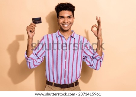 Photo of attractive young man show okey symbol hold credit card wear trendy striped formalwear clothes isolated on beige color background