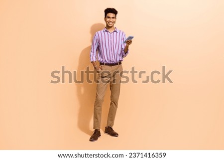 Full size photo of optimistic man dressed striped shirt brown trousers hold smartphone hand in pocket isolated on beige color background