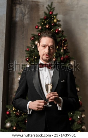 wealthy gentleman in suit with bow tie holding champagne glass near decorated Christmas tree