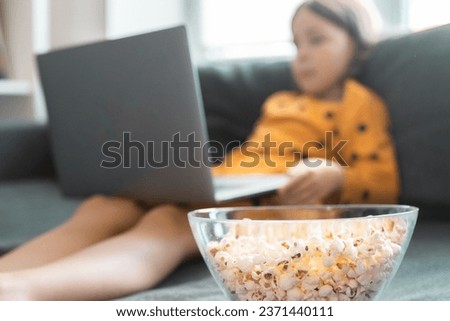 Little girl with laptop and popcorn sits on a grey couach. School girl watches educational video. Children and cartoons concept.