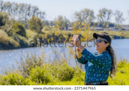 Smiling senior adult woman taking selfie with her mobile phone in countryside, river, trees and wild vegetation in blurred background, blue blouse, black cap and sunglasses, sunny autumn day