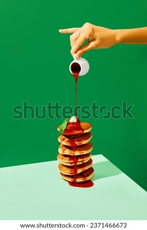 Food pop art photography. Close up. Female hand pouring sweet jam on delicious pancakes with butter over green background. Complementary colors. Complementary colors. Concept of food, art, cooking. Ad
