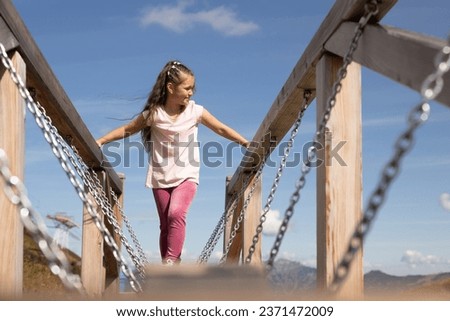 A girl walks on a beam suspended on metal chains on a mountain playground in the Austrian Alps Royalty-Free Stock Photo #2371472009