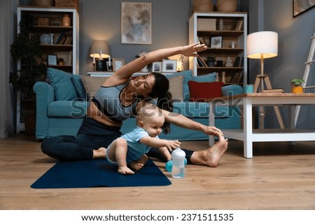 Young happy successful independent single mother is practicing yoga at home on a yoga mat while her baby is carelessly sitting on the floor and playing with a water bottle. Mom stretching muscles Royalty-Free Stock Photo #2371511535