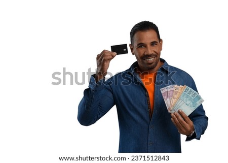 A man with Brazilian money in his hand holding a black card smiles looking at the food, wearing an orange T-shirt and a blue long-sleeved shirt on a white background.