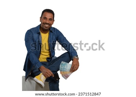 man holding brazilian money and a black credit card, sitting on a puff, wearing yellow and blue clothes, with yellow shoes on a white background