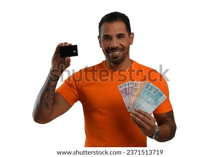 man holding a black credit card and Brazilian money, smiling, he wears a basic orange shirt on a white background