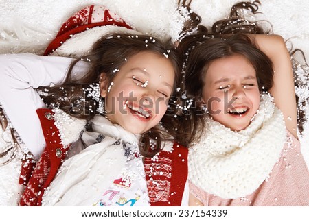 Two beautiful little girl with long hair in a white winter sweater lying in the snow under the Christmas tree and laugh gift for New Year Merry Christmas card