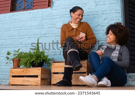 Happy brazilian adult daughter and mother sitting and relaxing and enjoying outside the house. Unity, happiness, affection, love, care concept. Royalty-Free Stock Photo #2371543645