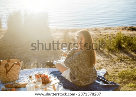 Beautiful young curly woman in warm cardigan on cozy autumn picnic by the lake with fruits, pastries and glass of white wine. Sun rays.