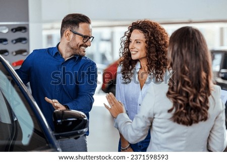A car salesman is showing new cars to a couple. Car sales business. Manager talking to couple, showing them new auto at dealership shop. Young family selecting vehicle, looking at modern automobile  Royalty-Free Stock Photo #2371598593