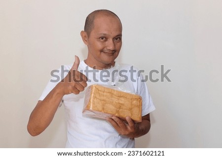 Expression of a bald Asian Indonesian wearing a white t-shirt against a white wall background. 
carrying a box of white bread which was still in plastic packaging.
