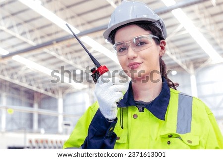 Female industrial engineer in hard hat uses laptop computer while standing in the heavy industry manufacturing factory. In the background various metalwork project parts, materials.