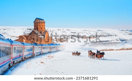 Red diesel train (East express) in motion at the snow covered railway - Horses pulling sleigh in winter - Ani Ruins, Ani is a ruined and  medieval Armenian city - Kars, Turkey Royalty-Free Stock Photo #2371681291