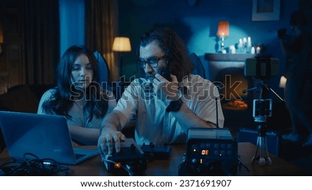 Medium-sized photo of a ghost hunter and a victim sitting at the table in a dark room. Another ghost hunter is trying to capture paranormal activity with an EMF detector.