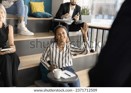 Young African American company intern listening to speaker lecture, raising hand for question to teacher. Diverse business group, multiethnic audience attending company training, lecture, presentation Royalty-Free Stock Photo #2371712509
