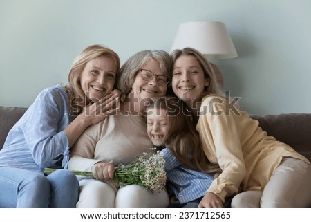 Happy joyful kid girl, mom, grandmother celebrating 8 march, great grandma birthday, holding bouquet of flowers, sitting close on sofa at home, hugging, smiling. Four family generations portrait