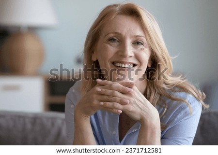 Cheerful blonde mature 60s lady home head shot portrait. Happy retired woman, pensioner, homeowner sitting on sofa with chin on hands, looking at camera with toothy smile, posing