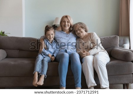 Three female generations sitting, relaxing on couch, enjoying meeting, leisure together. Happy mature grandma woman hugging beloved great grandmother and little granddaughter. Home family portrait