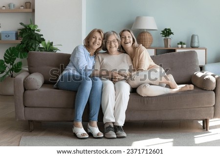 Happy united elder grandma, mature mom, young daughter woman sitting in couch in home interior, smiling, laughing, hugging the eldest woman with love, care, affection, looking at camera. Full length