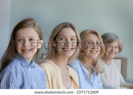 Cheerful pretty girls and women of four female generations posing indoors, standing in line, looking away with toothy smile. Kid, mom, grandmother, great grandma family home portrait