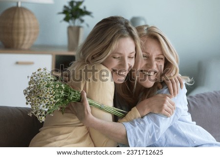 Cheerful excited loving young adult daughter congratulating mature mom on 8 march, mothers day, giving bunch of spring flowers, hugging mom with cheek touch. Senior woman enjoying happy motherhood