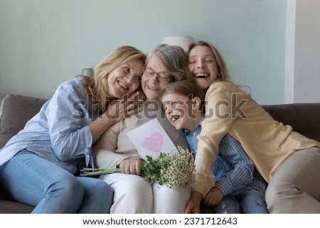Cheerful girls and women congratulating eldest great grandma on birthday, giving flowers, hand drawn greeting card with heart from kid. Four female family generation hugging, celebrating mothers day